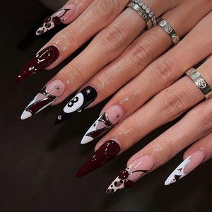 Puede incluir: Primer plano de uñas largas y puntiagudas con varios diseños. Algunas uñas son de color burdeos oscuro, otras tienen puntas blancas, detalles plateados y una uña negra con un diseño de bola ocho blanca. Varios anillos adornan los dedos.