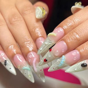 Puede incluir: Uñas acrílicas largas y puntiagudas con base rosa claro y puntas transparentes. Las uñas están decoradas con diseños 3D que incluyen mariquitas, gusanos y gotas de agua. Algunas uñas tienen un patrón de hojas blancas.