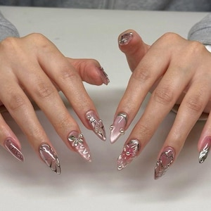 May include: Long, pointed acrylic nails with a clear, pink, and silver design. Each nail has a different embellishment, including silver accents, bows, and floral details. The nails are a light pink colour with a glossy finish.