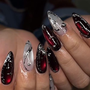 May include: A set of long, pointed acrylic nails. The nails alternate between a deep red and a translucent white, with silver embellishments. Some nails feature star and swirl designs, while others have small silver studs.