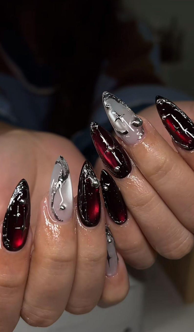 May include: A set of long, pointed acrylic nails. The nails alternate between a deep red and a translucent white, with silver embellishments. Some nails feature star and swirl designs, while others have small silver studs.