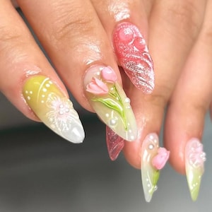 Puede incluir: Primer plano de una mano con uñas largas en forma de almendra. Las uñas están pintadas con una variedad de diseños, incluyendo tulipanes rosas en 3D, puntos blancos y patrones florales. Algunas uñas tienen un degradado de amarillo a blanco.