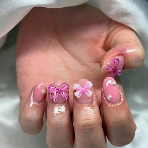 May include: A hand with pink nail polish and decorative bows and pearls. The nails are painted a light pink color, with some nails featuring 3D pink bows and small white pearls. The other nails have a clear bow or a pearl.