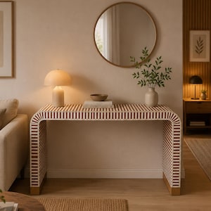 May include: A contemporary console table featuring a red and white striped design. The table has a curved shape and gold-coloured accents on the legs. A round mirror is mounted above, with a lamp and vase displayed on the table.