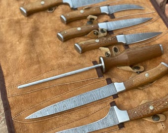 May include: A collection of various kitchen knives with wooden handles and patterned steel blades. The knives are displayed on a brown leather roll. The blades have a unique wood grain pattern. The handles are a light brown color.