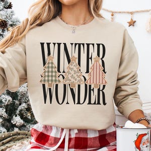 Könnte beinhalten: Ein beigefarbenes Sweatshirt mit Rundhalsausschnitt, schwarzem Schriftzug WINTER WONDER und drei stilisierten Weihnachtsbaumgrafiken. Die Bäume haben verschiedene Muster wie grünes Karomuster, Blumenmuster und rote Streifen mit einem Stern.