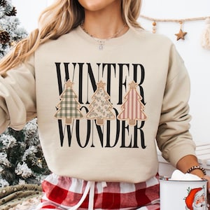 May include: A beige crewneck sweatshirt featuring the black text WINTER WONDER and three stylized Christmas tree graphics. The trees have different patterns: a green plaid, a floral print, and a red striped design, each topped with a star.