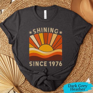Pode incluir: Uma t-shirt cinzenta escura mesclada com uma impress&atilde;o gr&aacute;fica de um sol a nascer sobre colinas e o texto "SHINING SINCE 1976". O design utiliza uma paleta de cores amarelo, laranja e castanho, com pequenos &iacute;cones de sol junto &agrave; palavra SHINING.