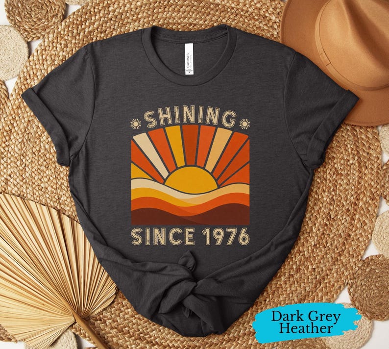 Pode incluir: Uma t-shirt cinzenta escura mesclada com uma impress&atilde;o gr&aacute;fica de um sol a nascer sobre colinas e o texto "SHINING SINCE 1976". O design utiliza uma paleta de cores amarelo, laranja e castanho, com pequenos &iacute;cones de sol junto &agrave; palavra SHINING.