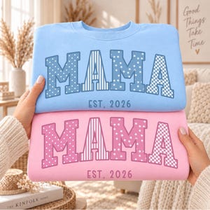 Könnte beinhalten: Ein hellblaues Sweatshirt und ein rosafarbenes Sweatshirt, jeweils mit dem Wort MAMA in großen Blockbuchstaben mit verschiedenen Mustern wie Punkten, Streifen, Sternen und Karos, sowie dem darunter gedruckten Text EST. 2026 in einfacher Schrift.