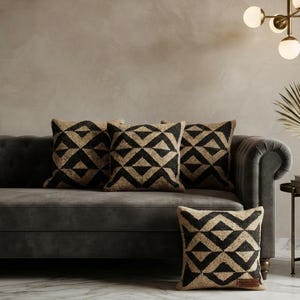 Könnte beinhalten: Vier quadratische Dekokissen mit einem sich wiederholenden geometrischen Rautenmuster in Schwarz und Beige. Die Kissen haben eine strukturierte Oberfläche und ein kleines rechteckiges Etikett an der unteren Ecke. Das Design besteht aus verbundenen Dreiecken.