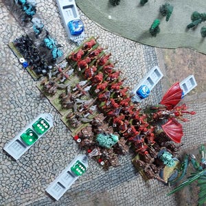 Peut inclure: Une scène de jeu de guerre sur table avec de nombreuses figurines miniatures, dont de l'infanterie peinte en rouge, un grand dragon rouge et un dragon vert. Plusieurs plateaux de mouvement en plastique gris portent les figurines et des dés.