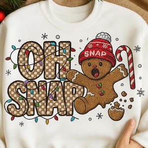 Op de afbeelding: Witte sweatshirt met een feestelijk "Oh Snap" ontwerp. De tekst is in een bruin geblokt patroon, met een peperkoekmannetje met een rode muts en een zuurstok. Kleurrijke lichtjes en sneeuwvlokken maken het kerstthema compleet.