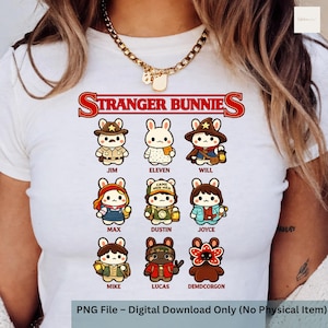 May include: White t-shirt featuring the text "STRANGER BUNNIES" in red, above cartoon bunny characters dressed as characters from the show. The characters include Jim, Eleven, Will, Max, Dustin, Joyce, Mike, Lucas, and Demdcorgon.