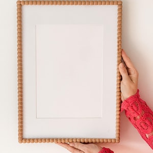 Puede incluir: Un marco de fotos rectangular con un paspartú blanco y un marco de madera con cuentas de color marrón claro. El marco lo sostiene una persona que lleva una manga de encaje roja. El marco está vacío, listo para una foto o una obra de arte.