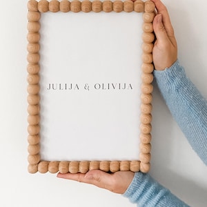 Puede incluir: Un marco rectangular de madera con borde de cuentas, sostenido por manos con un suéter azul claro. El marco tiene el texto "JULIJA & OLIVIJA".