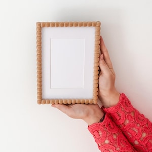 Op de afbeelding: Een rechthoekige houten fotolijst met een kralenrand. De lijst bevat een witte passe-partout en wordt door handen tegen een witte achtergrond gehouden. De persoon draagt een rode kanten mouw.