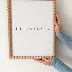 Puede incluir: Un marco rectangular de madera con borde de cuentas, sosteniendo un lienzo blanco. El texto "JULIJA & OLIVIJA" está centrado en el lienzo. El marco es sostenido por una persona que lleva un suéter azul claro.