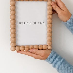 May include: A light-colored wooden picture frame with a beaded border is held against a white background. The frame has the text "JULIJA & OLIVIJA" in the center. The person holding the frame is wearing a light blue sweater.