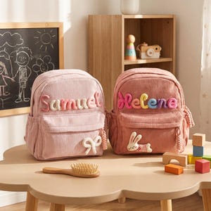 May include: Two corduroy backpacks in light pink and dusty rose. The light pink bag features the name Samuel in pink yarn and a white bow applique. The dusty rose bag features the name Helene in multicolored yarn and a white rabbit applique.