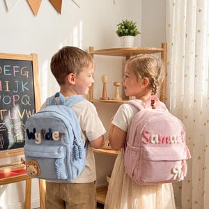 May include: A light blue corduroy backpack with the name Wylder and a small lion patch, and a light pink corduroy backpack with the name Samuel and a small bow patch. Both backpacks feature front zippered pockets and matching braided zipper pulls.