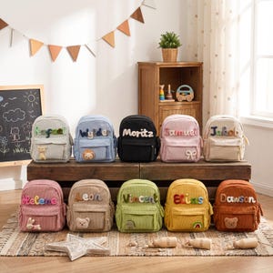 May include: A collection of ten small corduroy backpacks in various colors, each featuring a personalized name in colorful plush letters and a small animal or shape patch on the front pocket, arranged in two rows on a wooden surface.