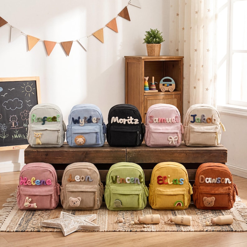 May include: A collection of ten small corduroy backpacks in various colors, each featuring a personalized name in colorful plush letters and a small animal or shape patch on the front pocket, arranged in two rows on a wooden surface.