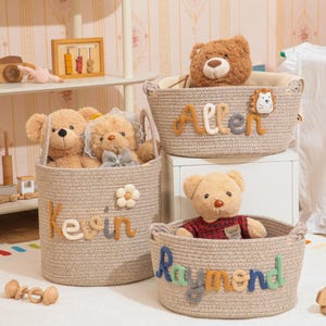 Puede incluir: Tres cestas de cuerda beige con osos de peluche dentro. Las cestas están etiquetadas con "Allen", "Kevin" y "Raymond" en letras coloridas. Un juguete de madera y otros elementos decorativos están en el fondo, creando un tema de guardería.