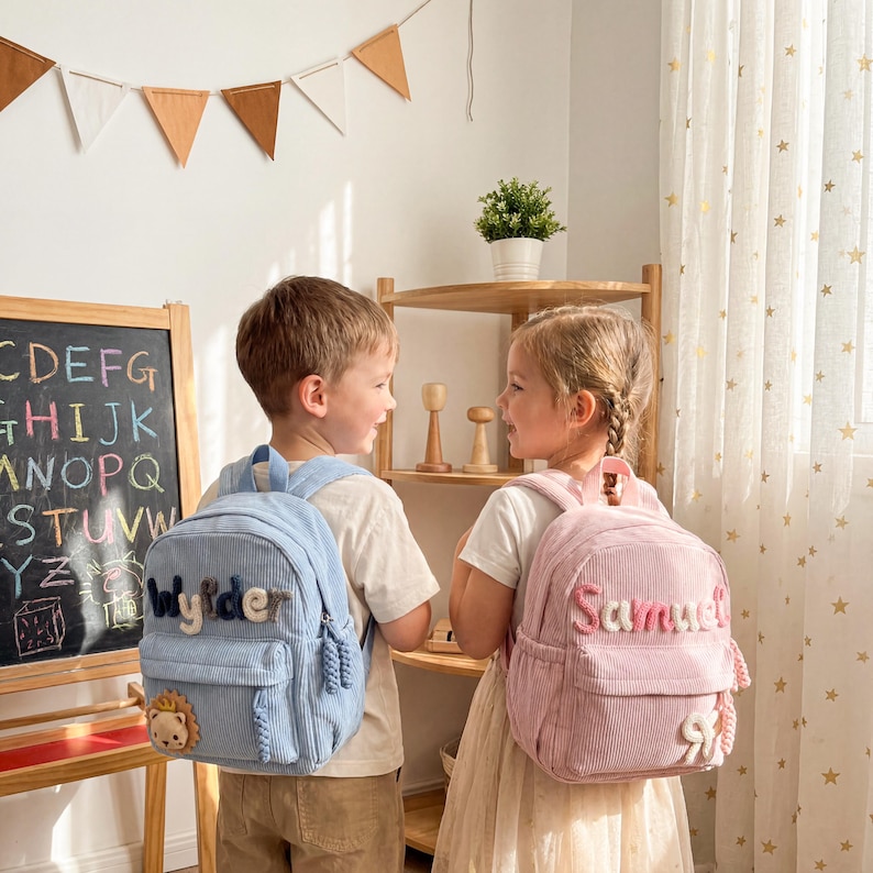 May include: A light blue corduroy backpack with the name Wylder and a small lion patch, and a light pink corduroy backpack with the name Samuel and a small bow patch. Both backpacks feature front zippered pockets and matching braided zipper pulls.