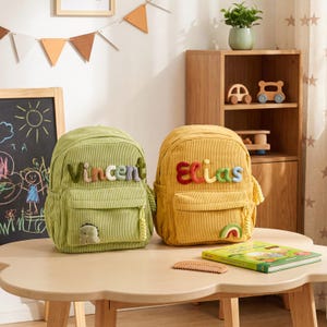 May include: Two corduroy backpacks in sage green and mustard yellow. The green bag features the name Vincent in textured letters and a small dinosaur patch. The yellow bag displays the name Elias in colorful letters and a rainbow patch.