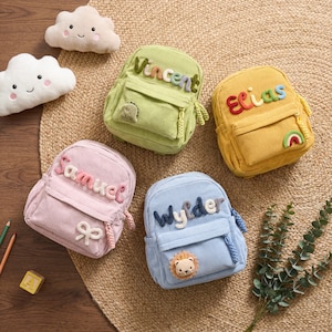 May include: Four corduroy backpacks in light green, yellow, pink, and light blue. Each bag features a personalized name in colorful, raised, textured letters, along with a small decorative patch like a dinosaur, rainbow, bow, or lion.
