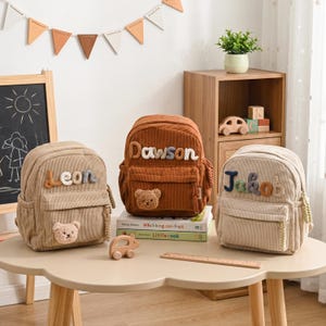 Puede incluir: Tres mochilas de pana en colores beige, marrón y crema. Cada bolsa cuenta con un bolsillo frontal con un parche de osito de peluche y un nombre bordado en letras coloridas y texturizadas: Leon, Dawson y Jakob.