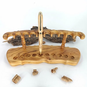 Può includere: Un portapenne in legno fatto a mano con una penna abbinata. Il supporto ha una base con più aperture circolari e una sezione superiore curva. La penna è di colore chiaro con dettagli dorati. Trucioli di legno sono sparsi.