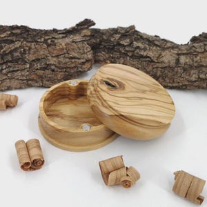 Puede incluir: Una caja redonda de madera clara con tapa, con un patrón de vetas prominente. La tapa está ligeramente abierta, revelando el interior. Pequeñas virutas de madera dispersas alrededor de la caja, con un trozo de corteza en el fondo.