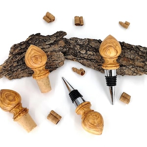 Può includere: Quattro tappi per bottiglie in legno con top decorativi, esposti su una superficie bianca con un pezzo di corteccia. I tappi hanno una finitura naturale venata e sono progettati per bottiglie di vino. Sono visibili componenti in sughero e metallo.