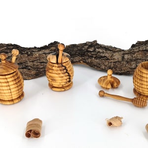 Può includere: Set di vasetti per miele in legno con un cucchiaio e due vasetti con coperchio, tutti realizzati in legno chiaro. I vasetti hanno un design a forma di alveare. Un pezzo di corteccia è sullo sfondo e trucioli di legno sono sparsi.