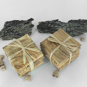 May include: Two stacks of square wooden coasters with a natural wood grain pattern, tied with raffia ribbon. The coasters are light brown and beige. Bark pieces are in the background.