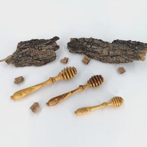 Può includere: Tre cucchiai di miele in legno con un design a coste sulla paletta e un manico decorativo. I cucchiai sono marrone chiaro con accenti marrone scuro. Piccoli pezzi di corteccia e trucioli di legno sono sparsi.