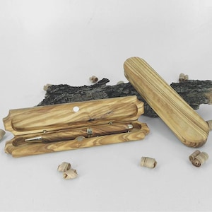 Può includere: Un set penna e astuccio in legno d'ulivo. La penna è all'interno di un astuccio aperto. L'astuccio è rettangolare con bordi arrotondati e chiusura magnetica. La penna ha dettagli argentati. L'astuccio è accanto a un astuccio chiuso e un pezzo di legno.
