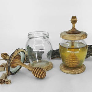 May include: Two glass honey jars with wooden lids and bases. One jar is filled with honey and has a wooden honey dipper. The other jar is empty. The lids and bases are made of light brown wood.