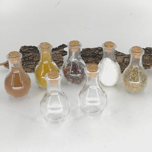 May include: A selection of small, clear glass spice jars with cork stoppers. The jars are filled with different spices, such as a brown powder, yellow powder, peppercorns, salt, and herbs. The jars are displayed on a white surface with a piece of bark in the background.