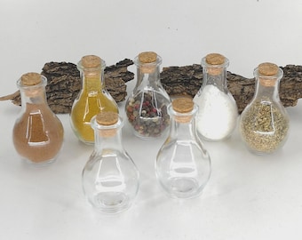 May include: A collection of small, clear glass spice jars with cork stoppers. The jars contain various spices, including a brown powder, yellow powder, peppercorns, salt, and herbs. The jars are arranged on a white surface with a piece of bark in the background.