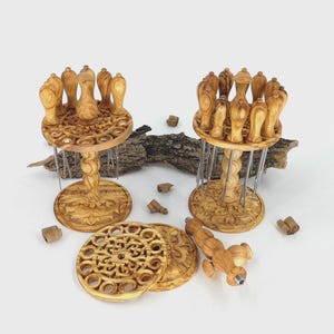 Op de afbeelding: Twee sets olijfhouten serveervorken met decoratieve bases en toppen, elk met meerdere vorken. Extra gesneden houten onderzetters en een serveervork zijn ook aanwezig. Het hout heeft een natuurlijke, lichtbruine kleur.