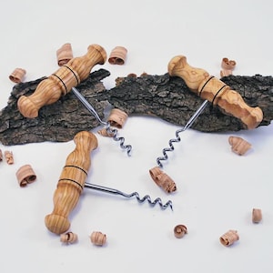 Pode incluir: Três saca-rolhas de madeira clara com espirais de aço inoxidável são exibidos em uma superfície branca. Os saca-rolhas são cercados por aparas de madeira e pedaços de casca de árvore, sugerindo um tema rústico.