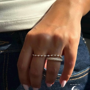 Puede incluir: Dos anillos de oro en un dedo. El anillo superior presenta una serie de pequeñas piedras claras de talla marquesa en una banda fina. El anillo inferior es una banda más ancha cubierta con un pavé de diminutas piedras brillantes. Texto: MOONLITJEWELLS.