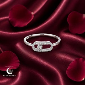 May include: A silver-toned ring featuring an elongated oval centerpiece encrusted with small clear stones. A single round clear stone is set within the center of the oval, designed to slide along the horizontal opening of the metal frame.