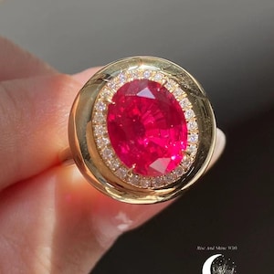 Può includere: Un anello d'oro con una grande pietra preziosa rossa brillante di forma ovale, circondata da un alone di piccole pietre chiare. L'anello ha una fascia d'oro semplice. L'anello è presentato su uno sfondo morbido.