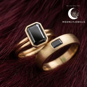 Könnte beinhalten: Zwei goldene Ringe mit schwarzen rechteckigen Steinen. Ein Ring hat einen großen Stein mit Goldfassung, der andere einen kleineren Stein. Die Ringe liegen auf einer dunkelroten, strukturierten Oberfläche. Der Markenname "MOONLITJEWELS" ist sichtbar.