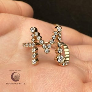 May include: A gold-coloured ring shaped like the letter "M", set with many small, clear gemstones. The ring is held in a hand, with the brand name "MOONLITJEWELLS" visible in the lower left corner.
