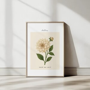 May include: A framed botanical print featuring a Cafe Au Lait dahlia illustration. The artwork showcases a cream-colored flower with green leaves and a bud, set against a beige background. The word "dahlias" is written above the flower, and "CAFE AU LAIT" is written below.